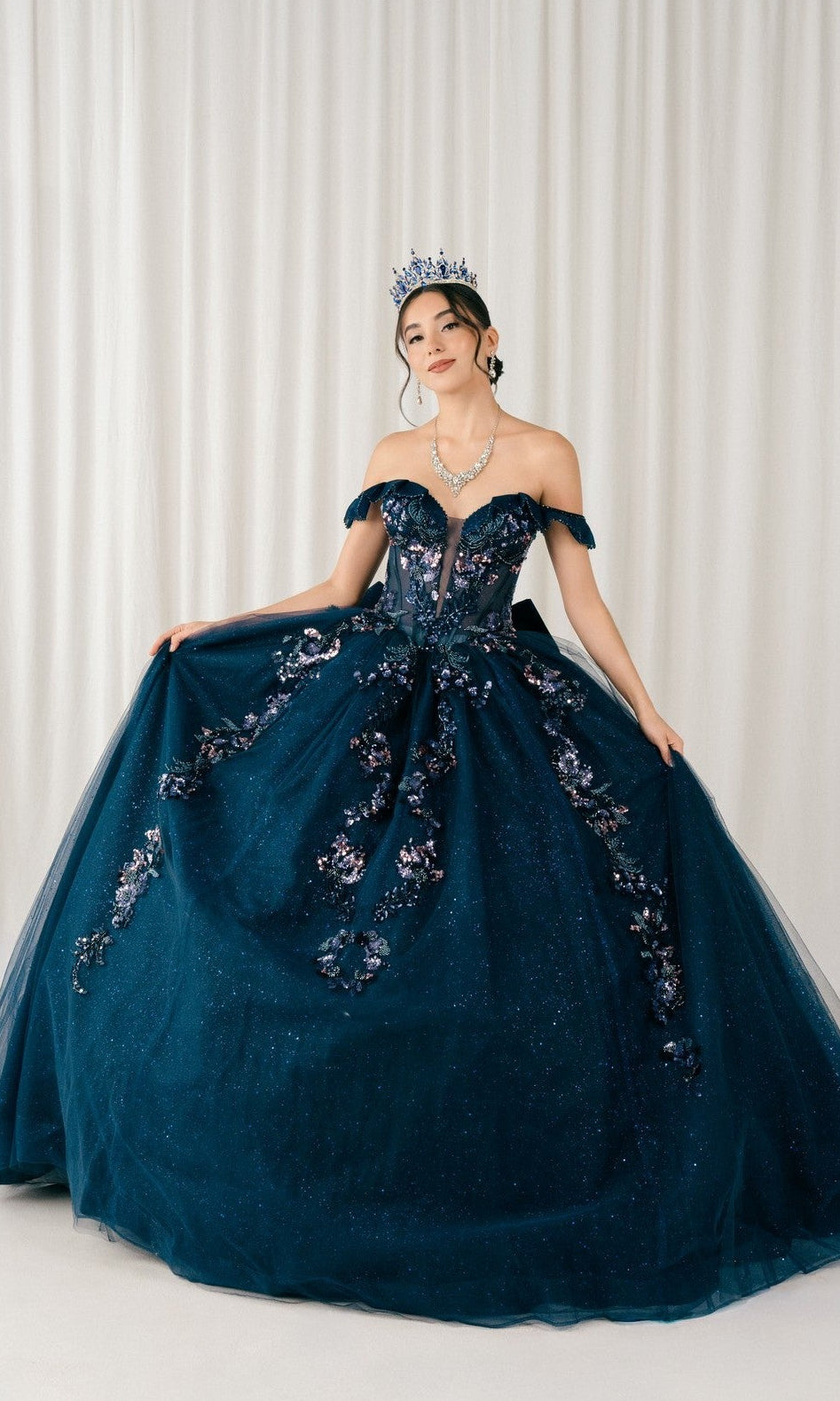 A woman stands before white curtains wearing the Quinceanera Dress 8440JQ by Cinderella Couture in navy, featuring off-the-shoulder floral embroidery and a matching tiara.